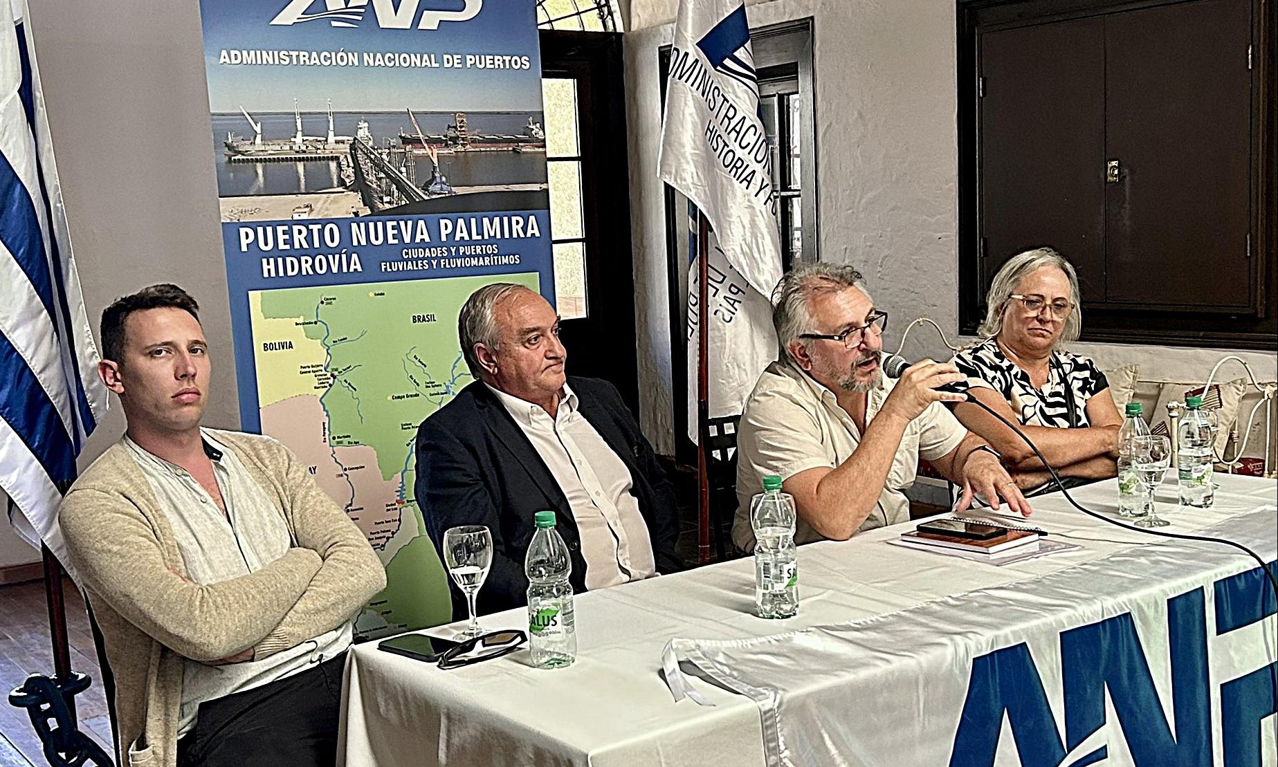 tres varones y una mujer sentados en una mesa frente a público, detrás cartel promocional del puerto de nueva palmira